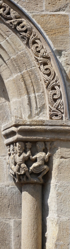 Right side of the interlaced tympanum. At the base of the arc, there is a sculpted capital with human figures holding hands, possibly dancing.