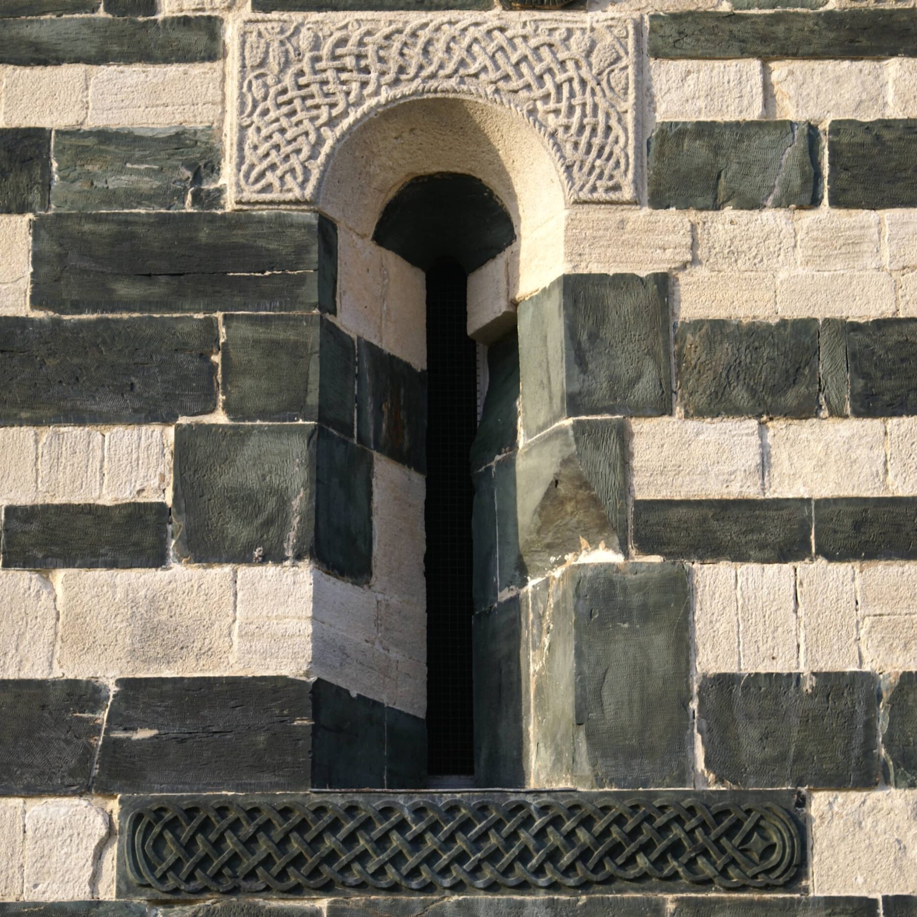 A loophole whose lintel and sill are carved with decorations: at the top, knotwork surrounded by vines on white limestone, at the bottom, a characteristic knotwork carved in green serpentinite stone.